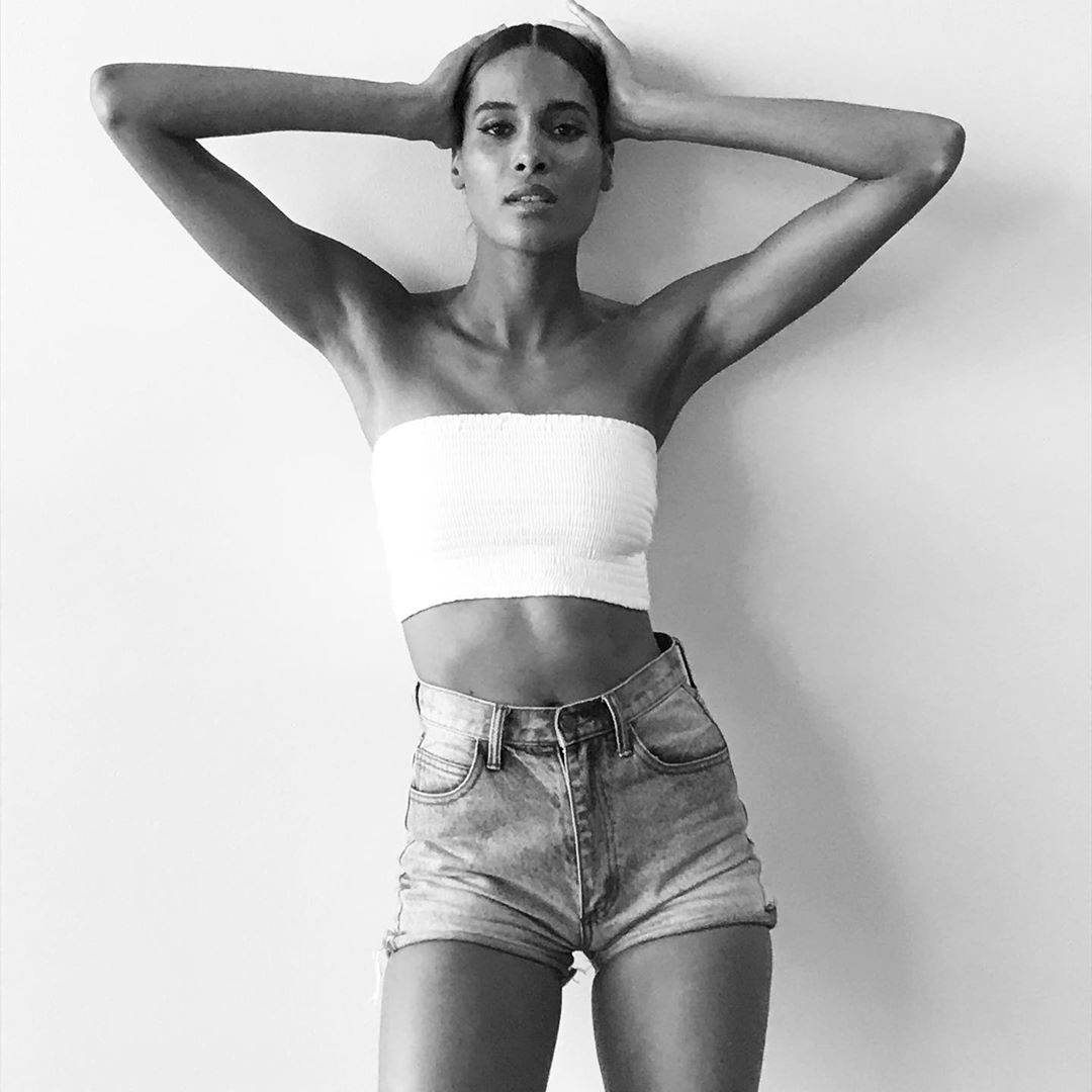 Black and white portrait of a woman in a tube top and denim shorts posing against a white wall, with her hands on her head.