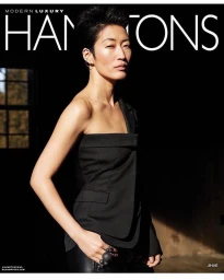 Woman with short black hair wearing a one-shoulder black top and dark trousers against a moody interior background.