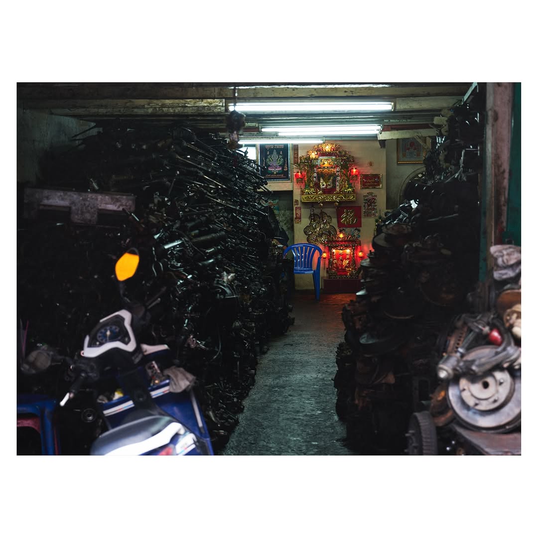 Indoor shot of stacked used motorcycle parts, filling the frame, with a glimpse of a shrine and blue chair in the background.