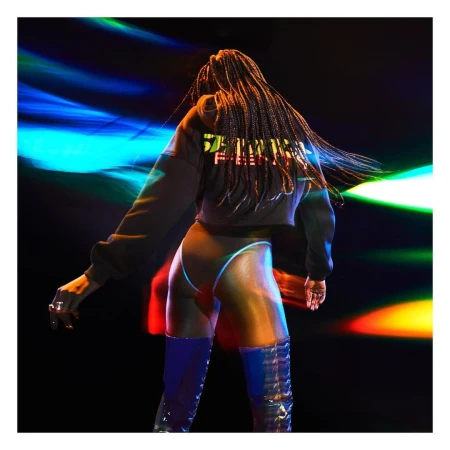 A woman with long braids wearing a black sweatshirt and blue thigh-high boots stands in front of a colorful blurred background.
