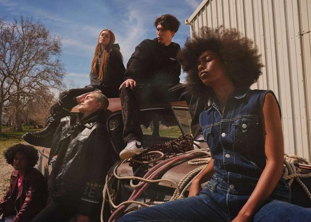 Group of five diverse young adults sit and pose on the hood of a rusty car outdoors on a sunny day, wearing trendy casual clothing.