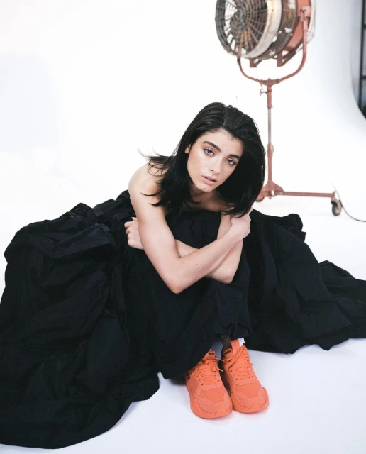 Studio portrait of a young woman sitting on the floor with a black dress and orange sneakers, posing with a fan in the background.