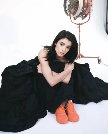 Studio portrait of a young woman sitting on the floor with a black dress and orange sneakers, posing with a fan in the background.