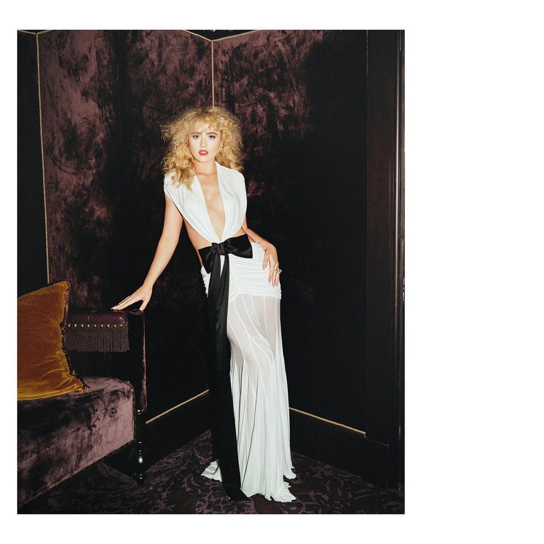 Woman in white gown with black bow posing near tufted sofa in dark interior. Fashion photography showcasing elegant evening wear.