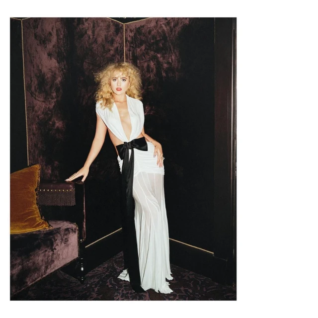 Woman in white gown with black bow posing near tufted sofa in dark interior. Fashion photography showcasing elegant evening wear.