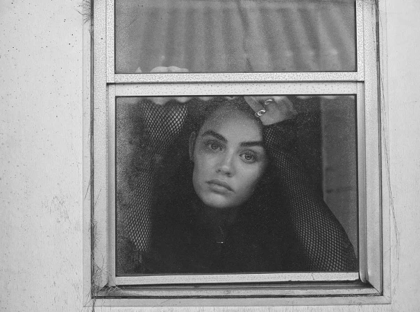 Black and white portrait of a woman looking out through a window pane wearing a mesh top shot by Dennis Leupold