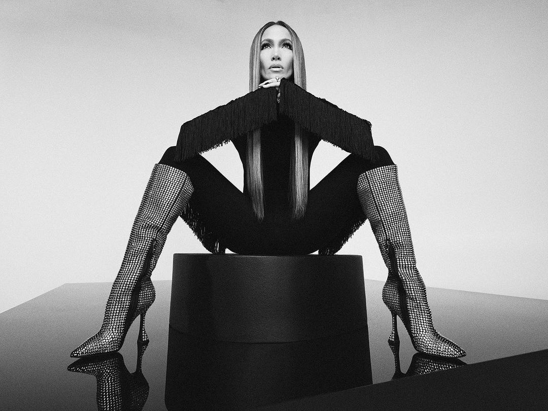 Jennifer Lopez seated on a dark pedestal wearing embellished rhinestone boots and fringed sleeves in a monochrome studio portrait.
