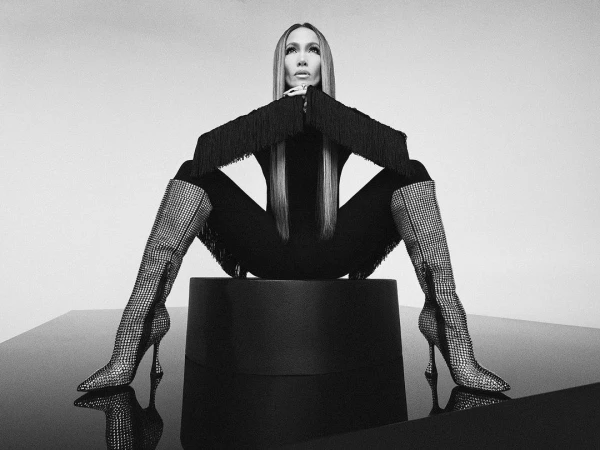 Jennifer Lopez seated on a dark pedestal wearing embellished rhinestone boots and fringed sleeves in a monochrome studio portrait.