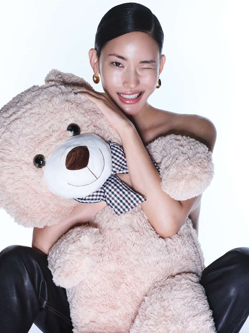 Smiling Asian woman with wet-look hair poses with a large teddy bear, wearing black pants and gold earrings against white backdrop.