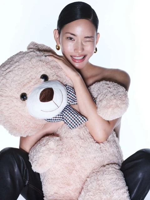 Smiling Asian woman with wet-look hair poses with a large teddy bear, wearing black pants and gold earrings against white backdrop.