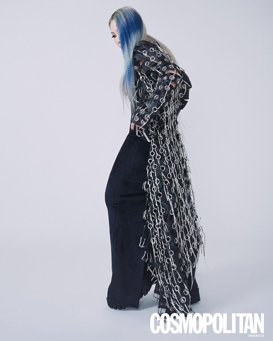 A slender adult female model poses in loose black pants and a long black top decorated with silver rings, against a white backdrop.