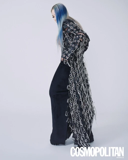 A slender adult female model poses in loose black pants and a long black top decorated with silver rings, against a white backdrop.