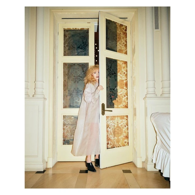 A woman in a light pink robe poses in a doorway in an ornate interior with off-white walls and decorative translucent panels in the doors.