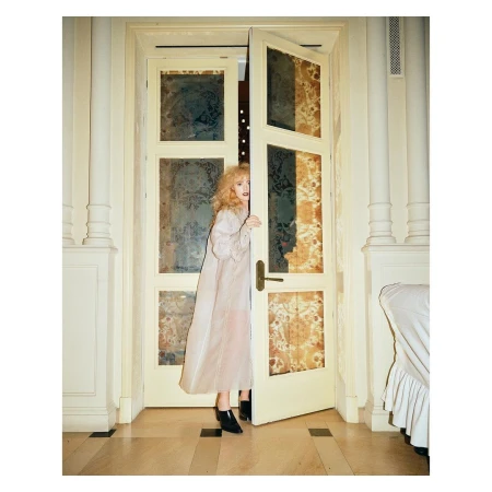 A woman in a light pink robe poses in a doorway in an ornate interior with off-white walls and decorative translucent panels in the doors.