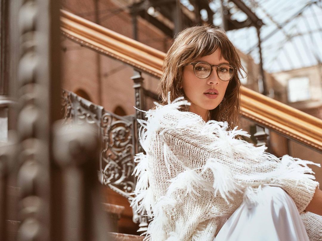 Portrait of a young woman with brown hair wearing glasses and a white sweater, posing indoors with iron railings in the background.