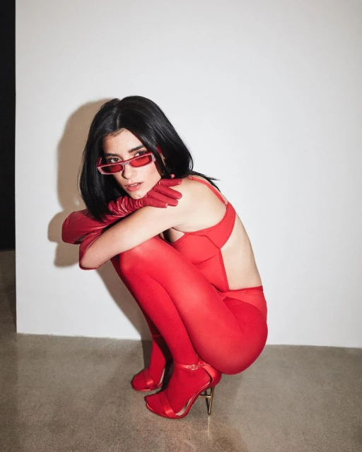 A young brunette woman with red accessories poses indoors, wearing matching sunglasses, gloves, heels, and bodysuit against a white backdrop.