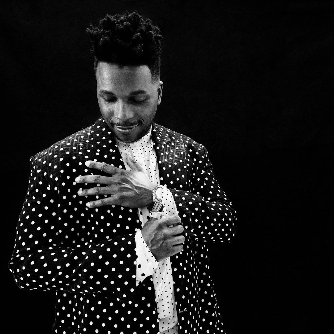 Black and white portrait of a stylish man wearing a polka dot blazer and adjusting his sleeve, set against a dark background.
