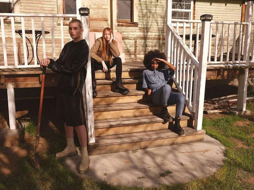 Three people on the porch stairs of a weathered house: one standing with a shovel, two seated on steps in casual outdoor fashion.