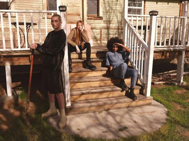 Three people on the porch stairs of a weathered house: one standing with a shovel, two seated on steps in casual outdoor fashion.