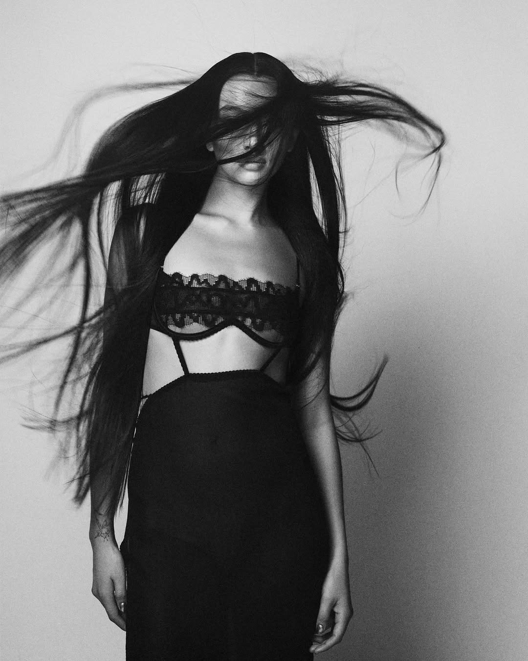 Black and white portrait of a woman with long dark hair blowing across her face wearing a dark lace lingerie top.