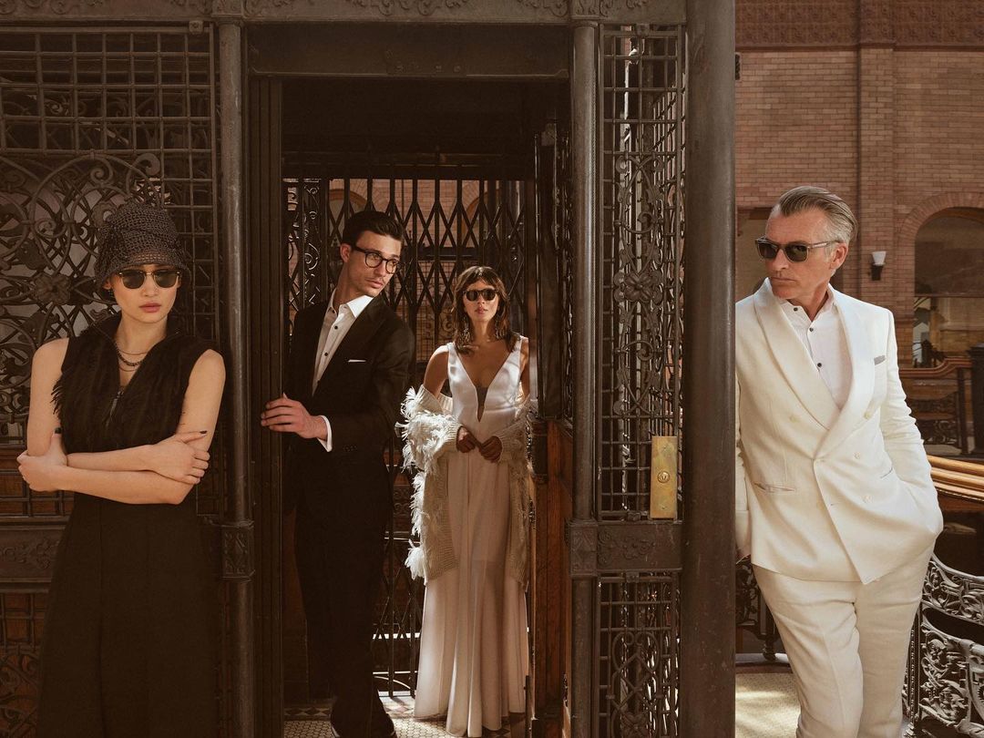 Four people posing inside a vintage elevator. An Asian woman, a man in glasses, a woman in white, and an older man in a white suit.