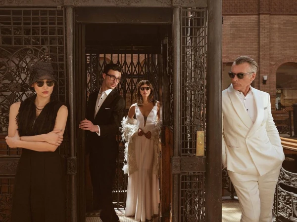 Four people posing inside a vintage elevator. An Asian woman, a man in glasses, a woman in white, and an older man in a white suit.