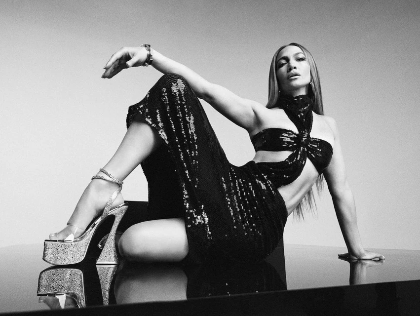 Jennifer Lopez poses in a black sequin dress and platform heels against a gradient grey backdrop in a studio-style setting.