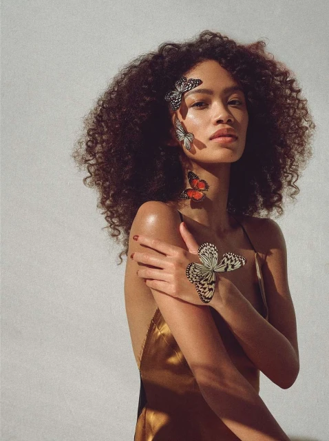 A woman with curly hair adorns butterflies on her face and body, wearing a golden dress, posing against a neutral background in soft lighting.