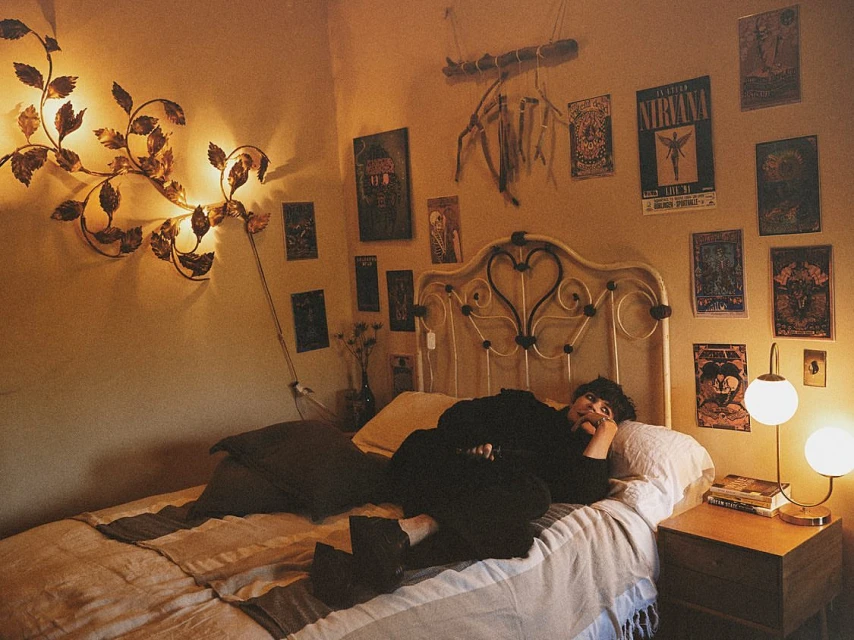 A person relaxes on a bed in a bedroom with posters, decorative lighting, and bedside table, creating a warm and inviting atmosphere.