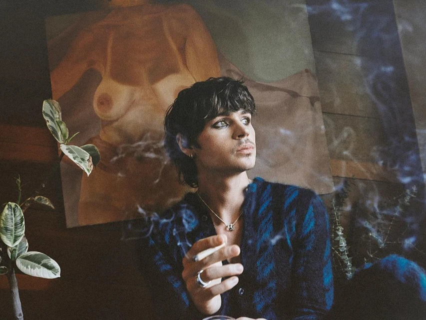 Adult man with dark hair and eye makeup poses indoors with a lit cigarette. A plant and an art print are visible in the background.