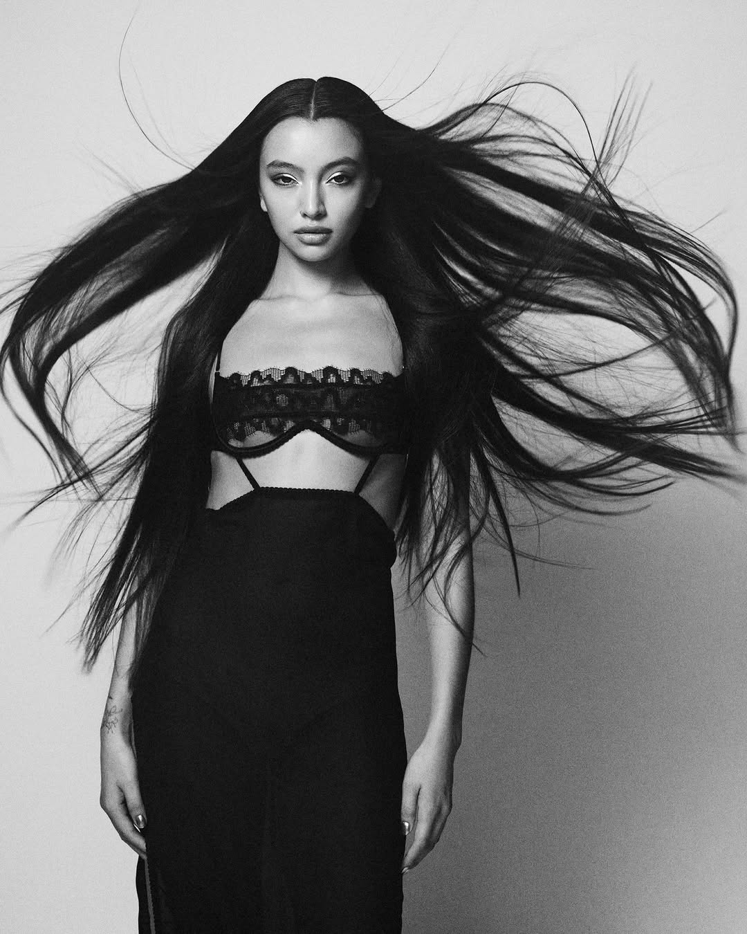 Striking black and white portrait of a woman with long hair, wearing a stylish black lace bra and dress, against a plain backdrop.