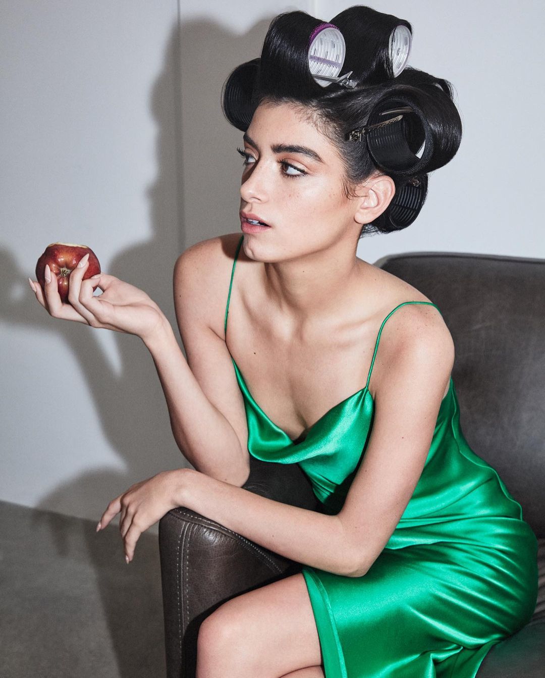 Adult woman with hair rollers poses with a red apple in a photo studio wearing a green silk dress. Beauty and fashion portrait.