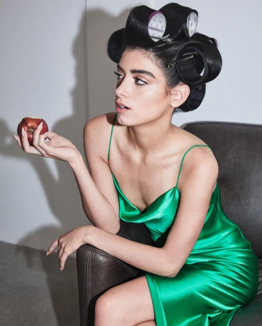 Adult woman with hair rollers poses with a red apple in a photo studio wearing a green silk dress. Beauty and fashion portrait.