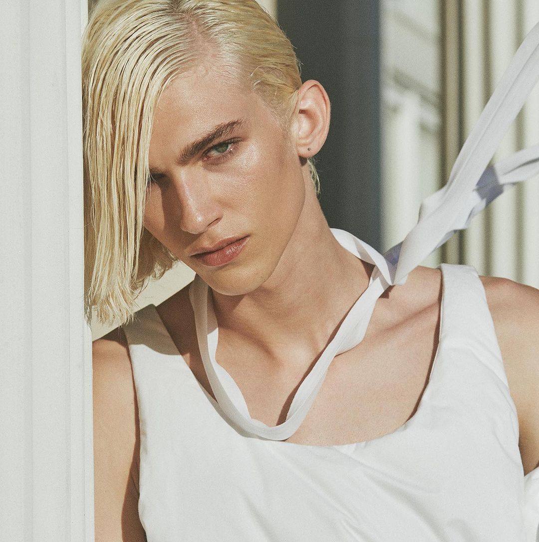 Close-up portrait of a young adult male with blonde hair, wearing a white tank top, leaning against a white pillar outdoors.