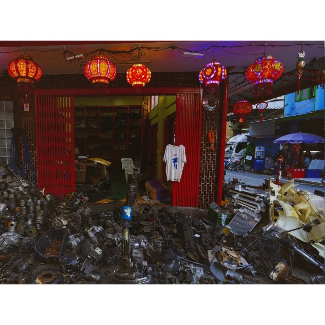 Outdoor salvage shop with salvaged car parts piled high, hanging red lanterns, and a white t-shirt hanging in doorway.