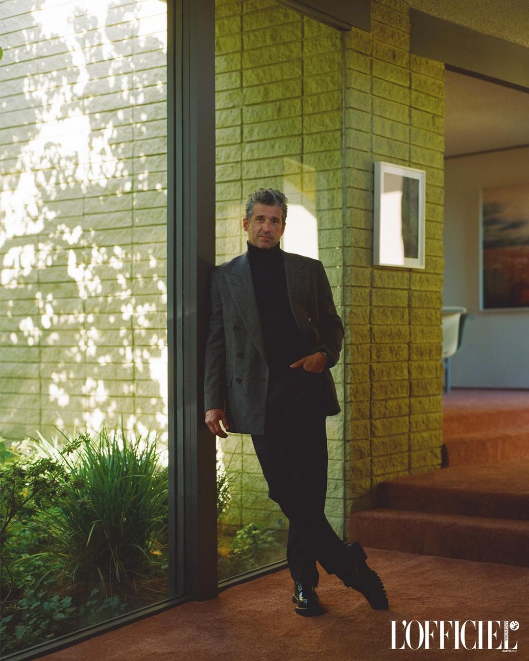 Mature man in a suit leans against a window in a modern home, with sunlight streaming in and L'Officiel logo at the bottom right.