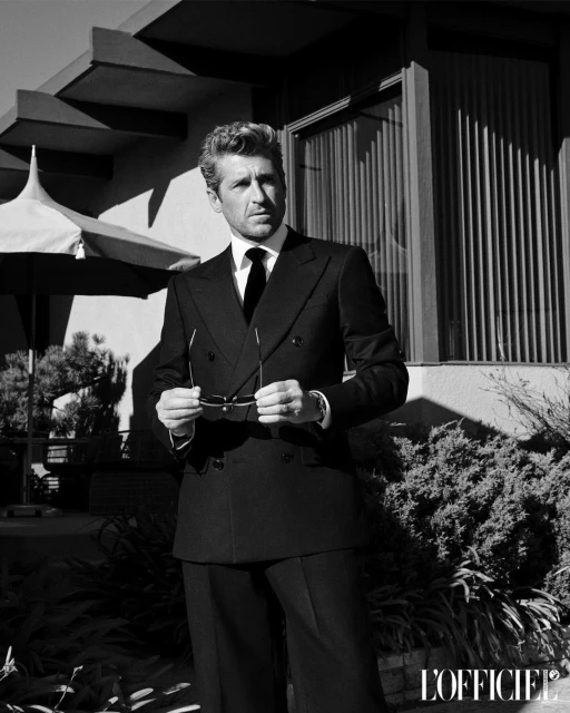 Patrick Dempsey poses in a double-breasted suit against a modern architectural backdrop, his gaze directed, holding sunglasses in black and white.