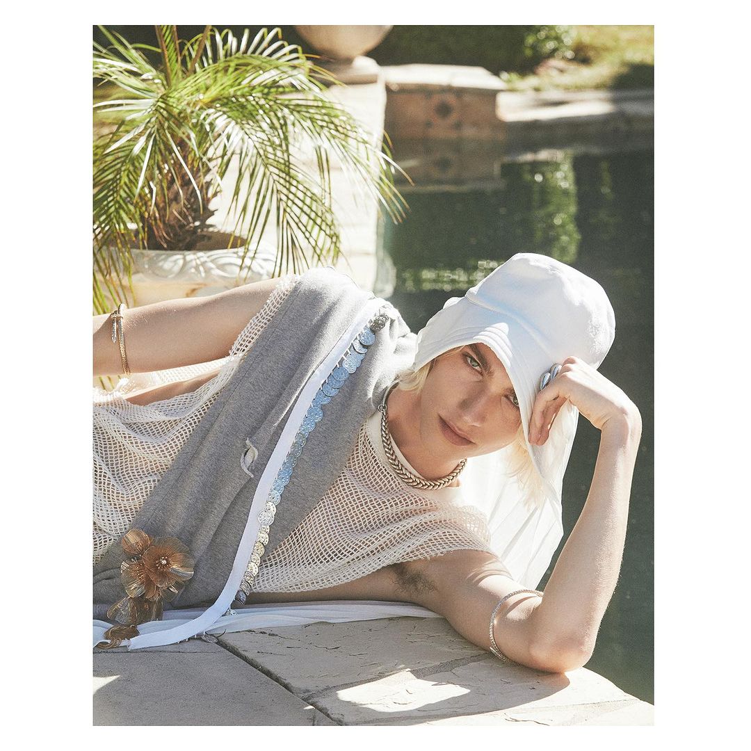 A light-skinned person wearing a white hat and draped in gray, posing on a stone ledge next to a green, leafy palm tree and body of water.