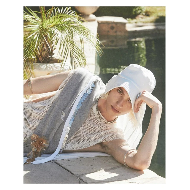 A light-skinned person wearing a white hat and draped in gray, posing on a stone ledge next to a green, leafy palm tree and body of water.
