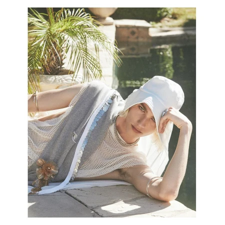 A light-skinned person wearing a white hat and draped in gray, posing on a stone ledge next to a green, leafy palm tree and body of water.