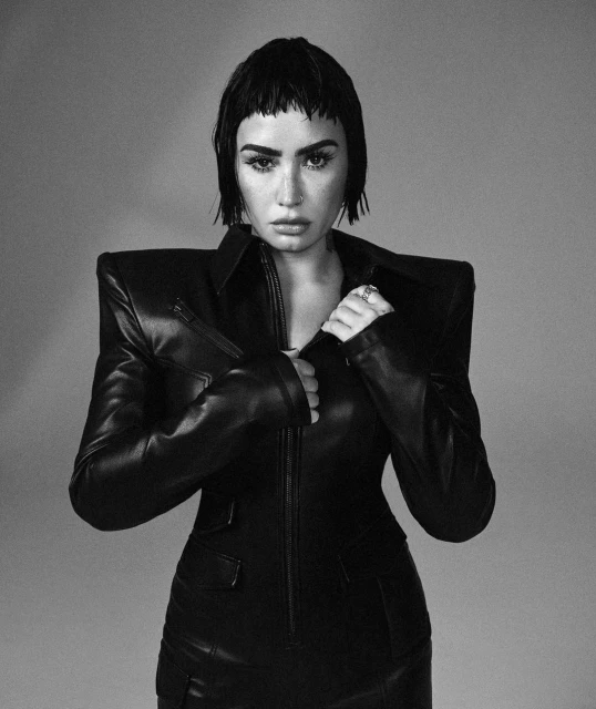 Black and white portrait of Demi Lovato in a dark leather jacket, looking at the camera with a neutral expression against a grey background.