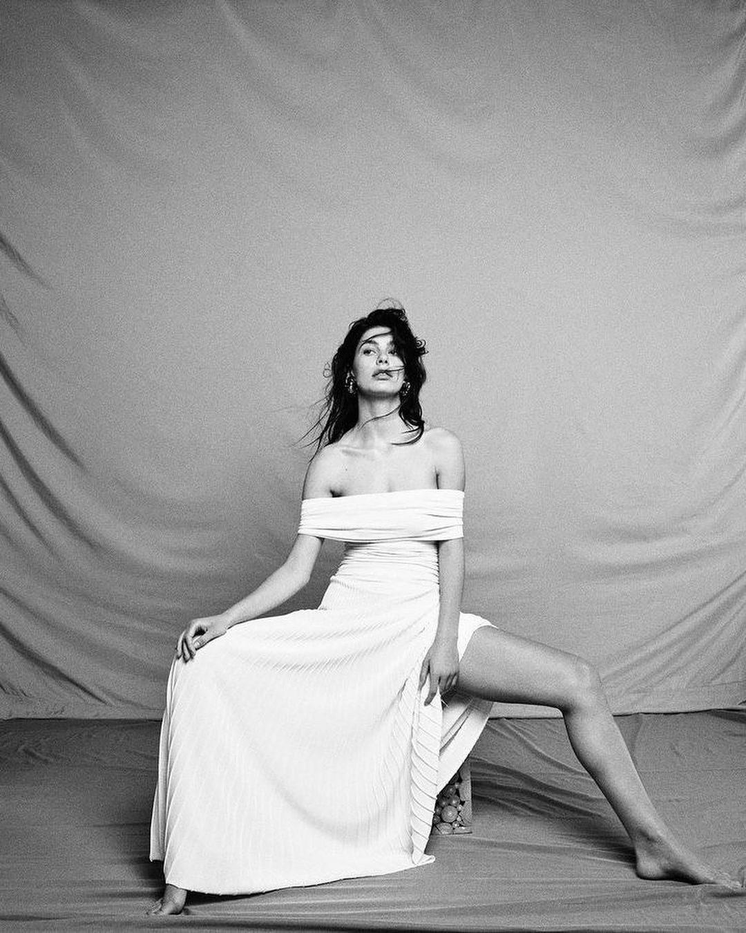 A woman poses in a white off-the-shoulder dress, seated with one leg extended in front of a draped fabric backdrop in black and white.