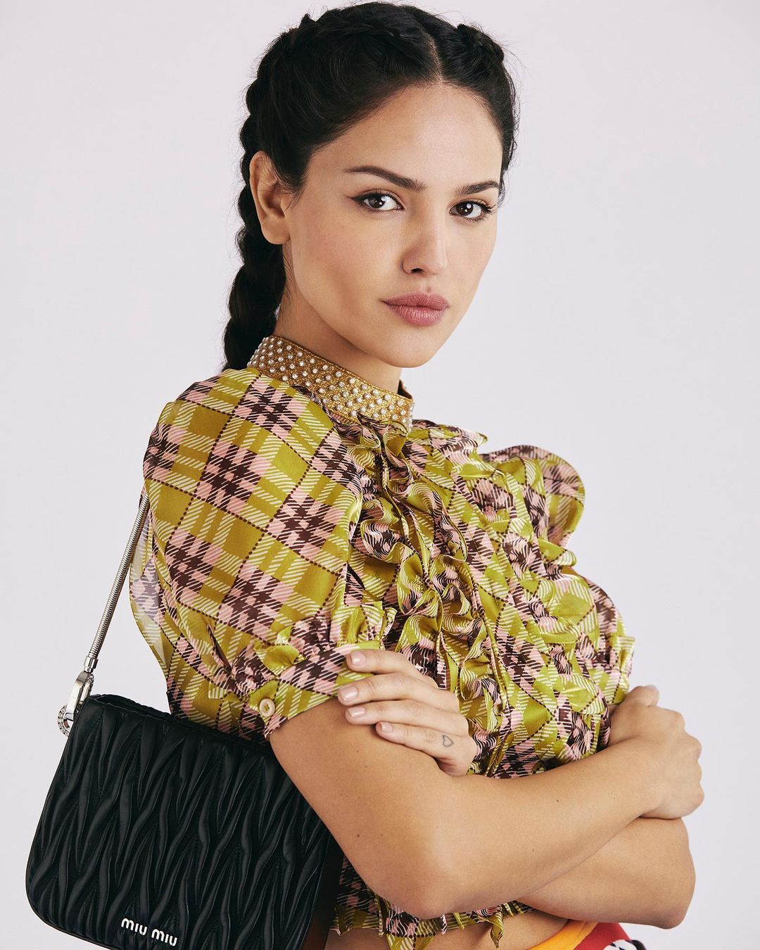 Brunette woman in plaid ruffled blouse with braided hair poses with black Miu Miu handbag in front of a white studio backdrop.