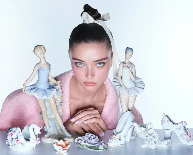 Young woman poses with porcelain ballerina figurines and swan planter on a white surface in a soft, diffused light, wearing a pink jacket.