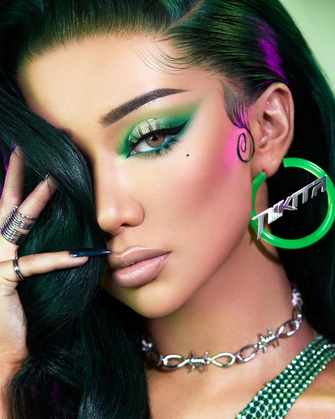 Close-up studio portrait of a woman with green hair, eye makeup, and jewelry against a gradient green and pink backdrop.
