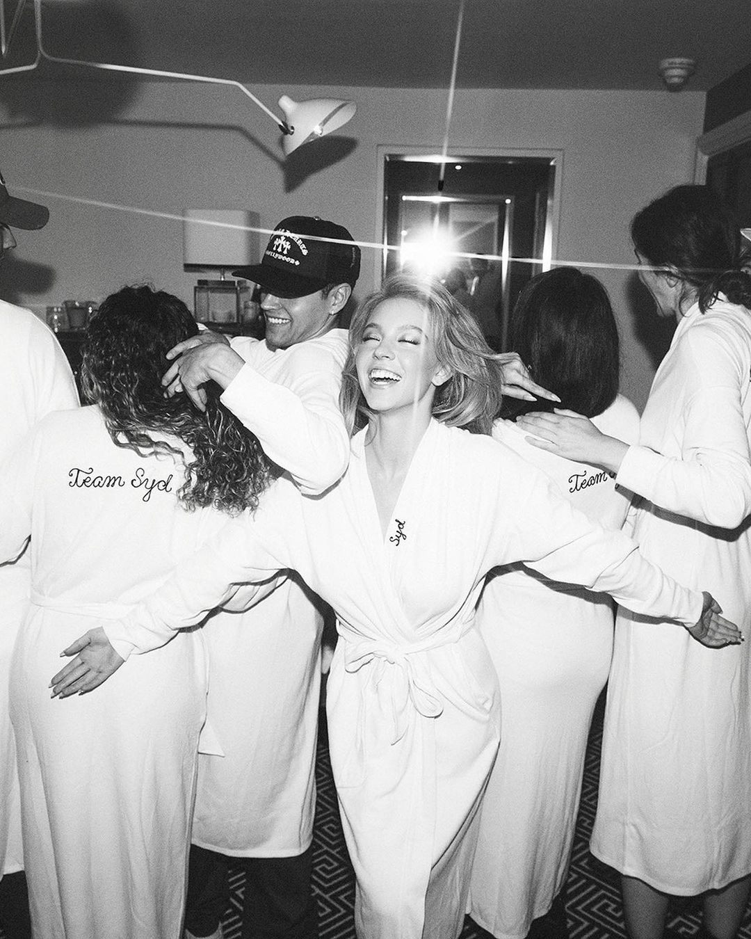 Black and white photo of Sydney Sweeney in a white robe, surrounded by her team in matching robes, celebrating in an indoor setting.