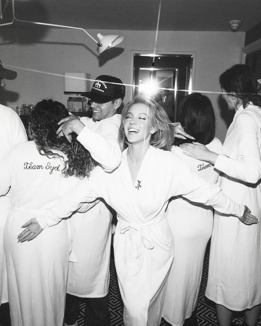 Black and white photo of Sydney Sweeney in a white robe, surrounded by her team in matching robes, celebrating in an indoor setting.