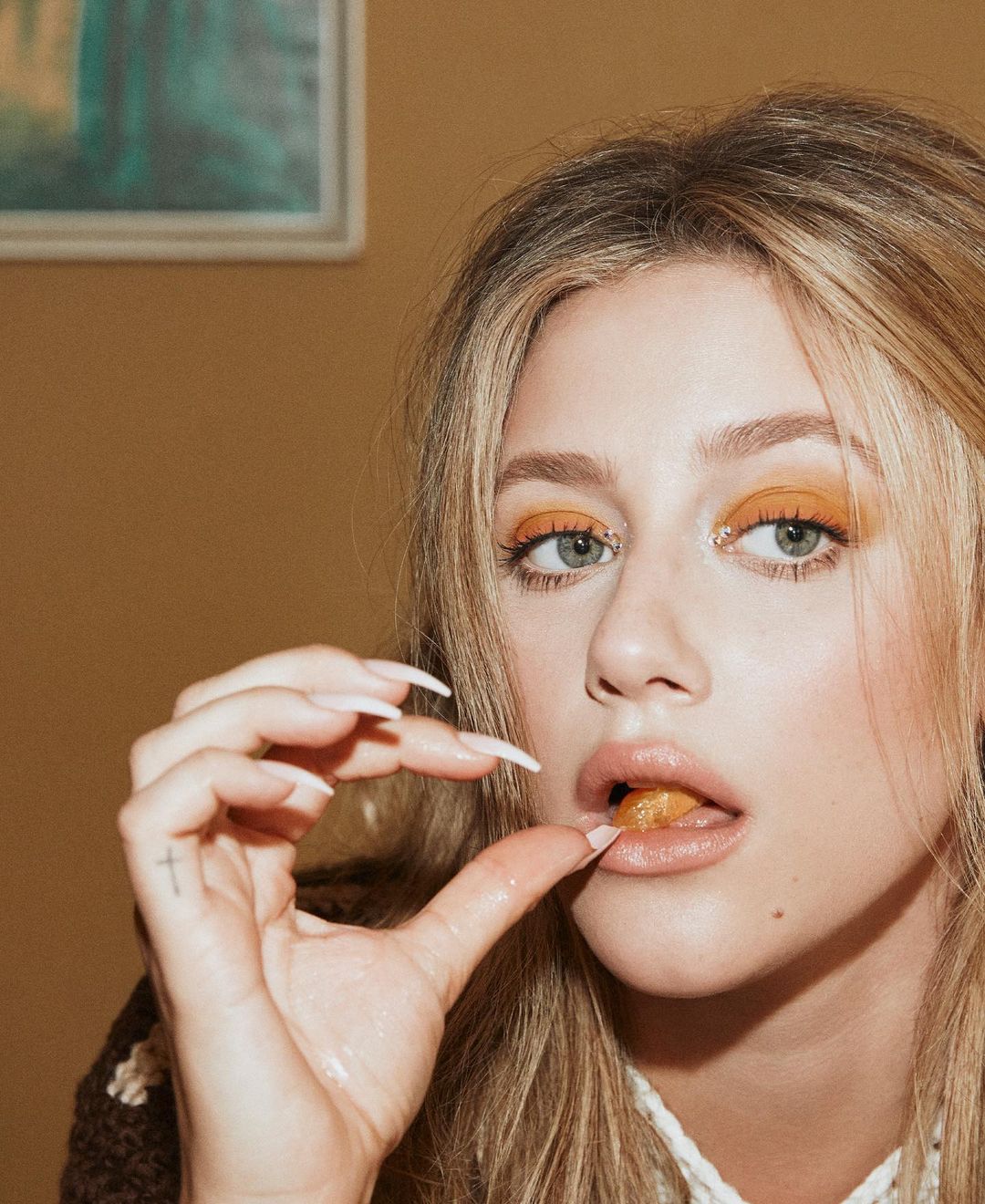 Close-up portrait of an adult woman eating, featuring her blonde hair, orange eyeshadow, and long nails against a solid tan background.