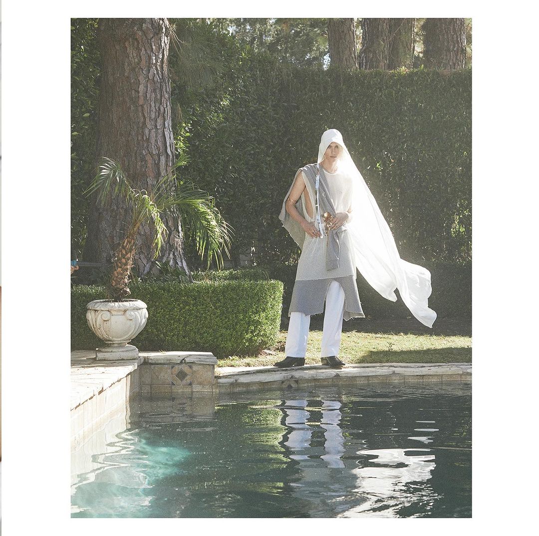 Model wearing a white hooded tunic and trousers standing by a swimming pool outdoors with trees in the background