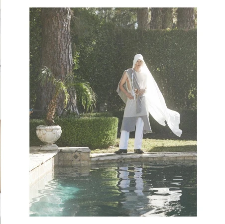 Model wearing a white hooded tunic and trousers standing by a swimming pool outdoors with trees in the background
